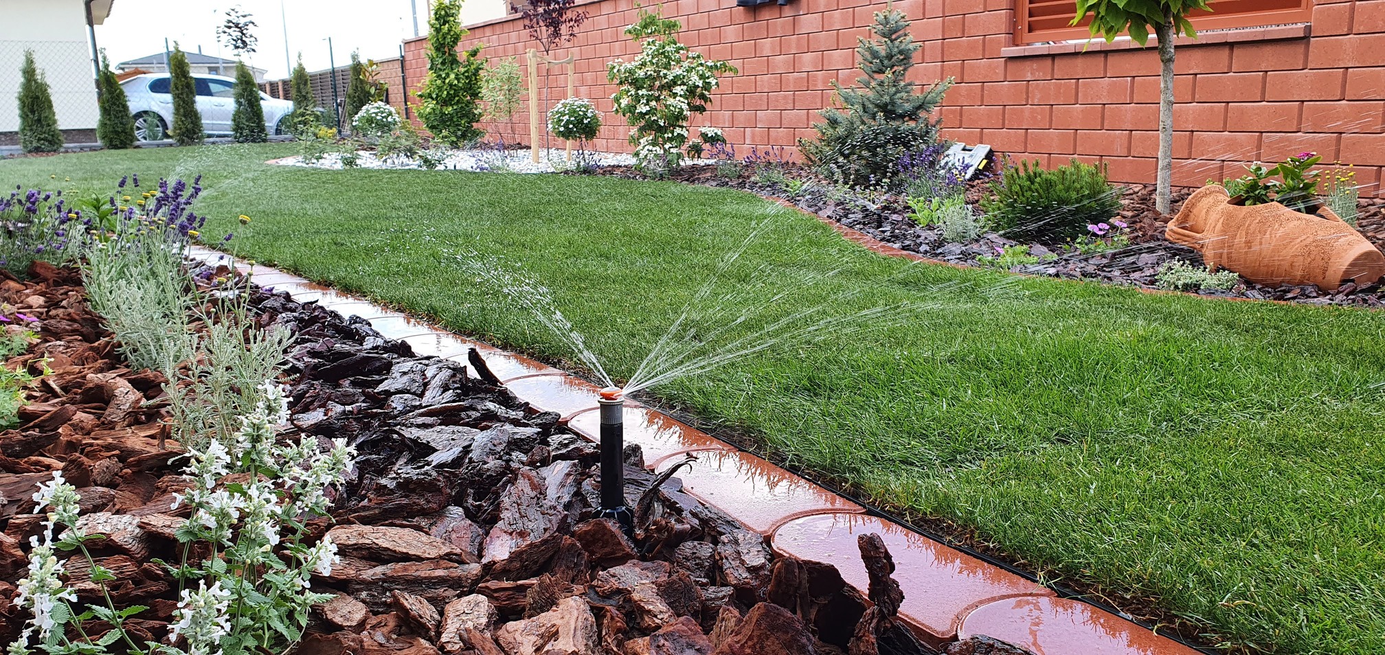 Výsuvný postrekovač zavlažujúci trávnik pri okrasnom záhone a tehlovom múre